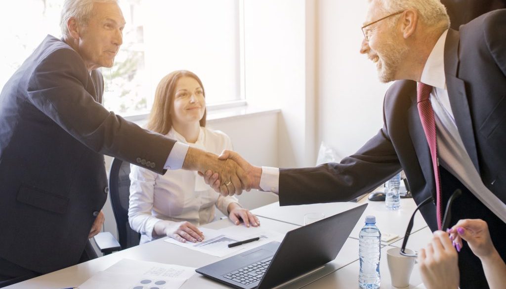 smiling-woman-looking-two-senior-businessman-shaking-hands