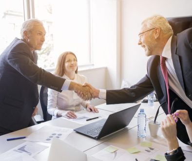 smiling-woman-looking-two-senior-businessman-shaking-hands